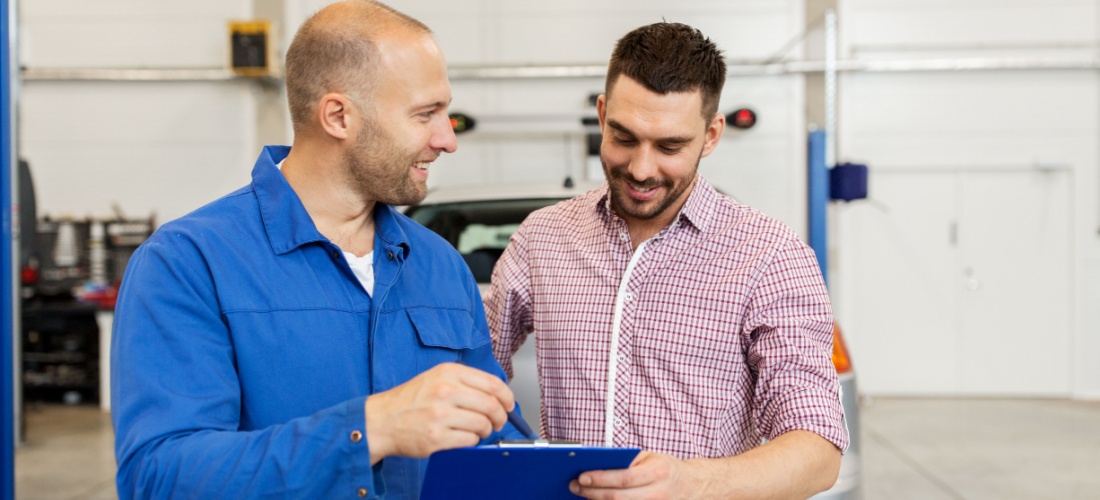 Un mecánico con un coche gris detrás le enseña a un cliente unos papeles.