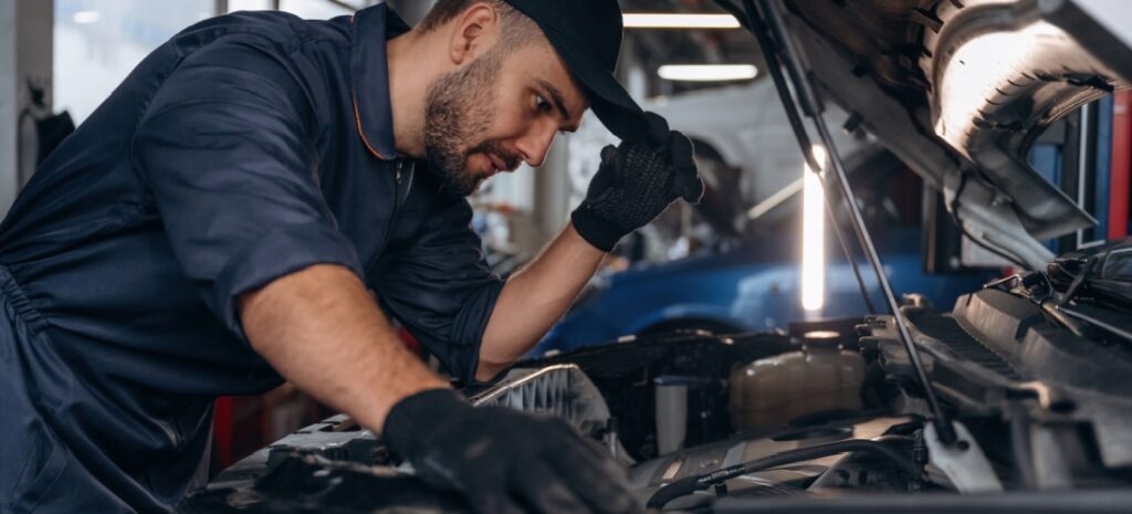 Mecánico se toca la gorra con gesto de preocupación mientras revisa el capó de un coche estropeado en el taller