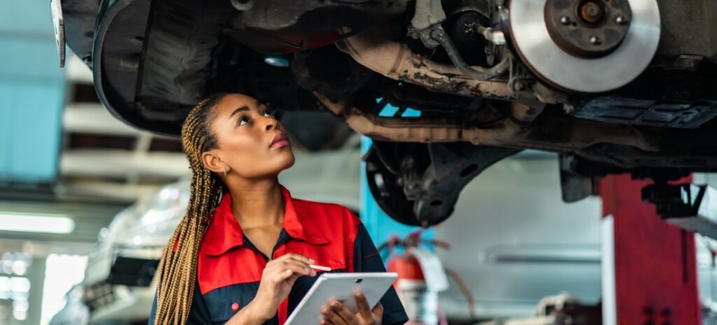 Mujer de color con trenzas rubias y tablet blanca en la mano, revisa la parte baja de un coche.